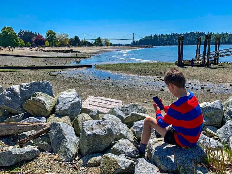 boy in red and blue striped shirt sitting on ambleside beach in vancouver looking at his cell phone